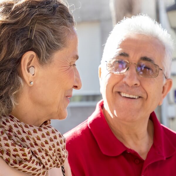 Smiling senior couple outdoors with a woman wearing TU SHOP micro hearing aids.