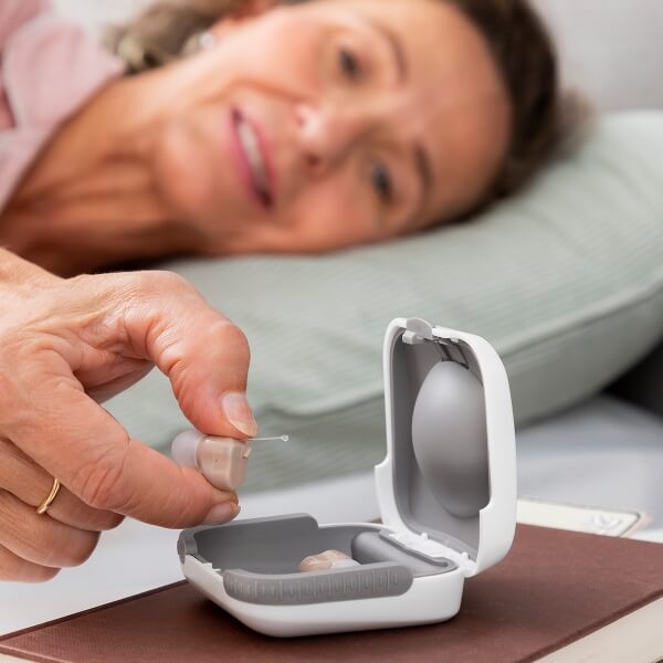 A woman holding a small TU SHOP micro hearing aid from its charging case.